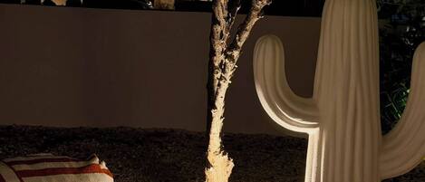 A cactus sculpture, a striped pillow, and a tree with a lit base in a nighttime outdoor setting.