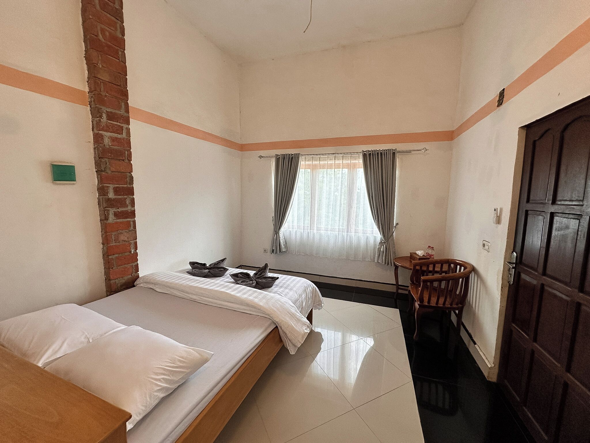Une petite chambre avec un lit simple, un mur en briques, une fenêtre avec des rideaux, une porte en bois et une petite table avec une chaise.