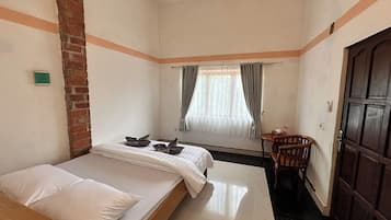 Une petite chambre avec un lit simple, un mur en briques, une fenêtre avec des rideaux, une porte en bois et une petite table avec une chaise.