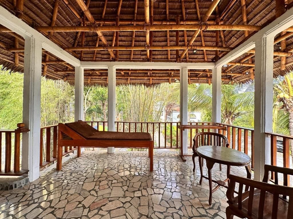 A covered outdoor area with a stone floor, wooden furniture, and a view of greenery.