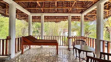 A covered outdoor area with a stone floor, wooden furniture, and a view of greenery.