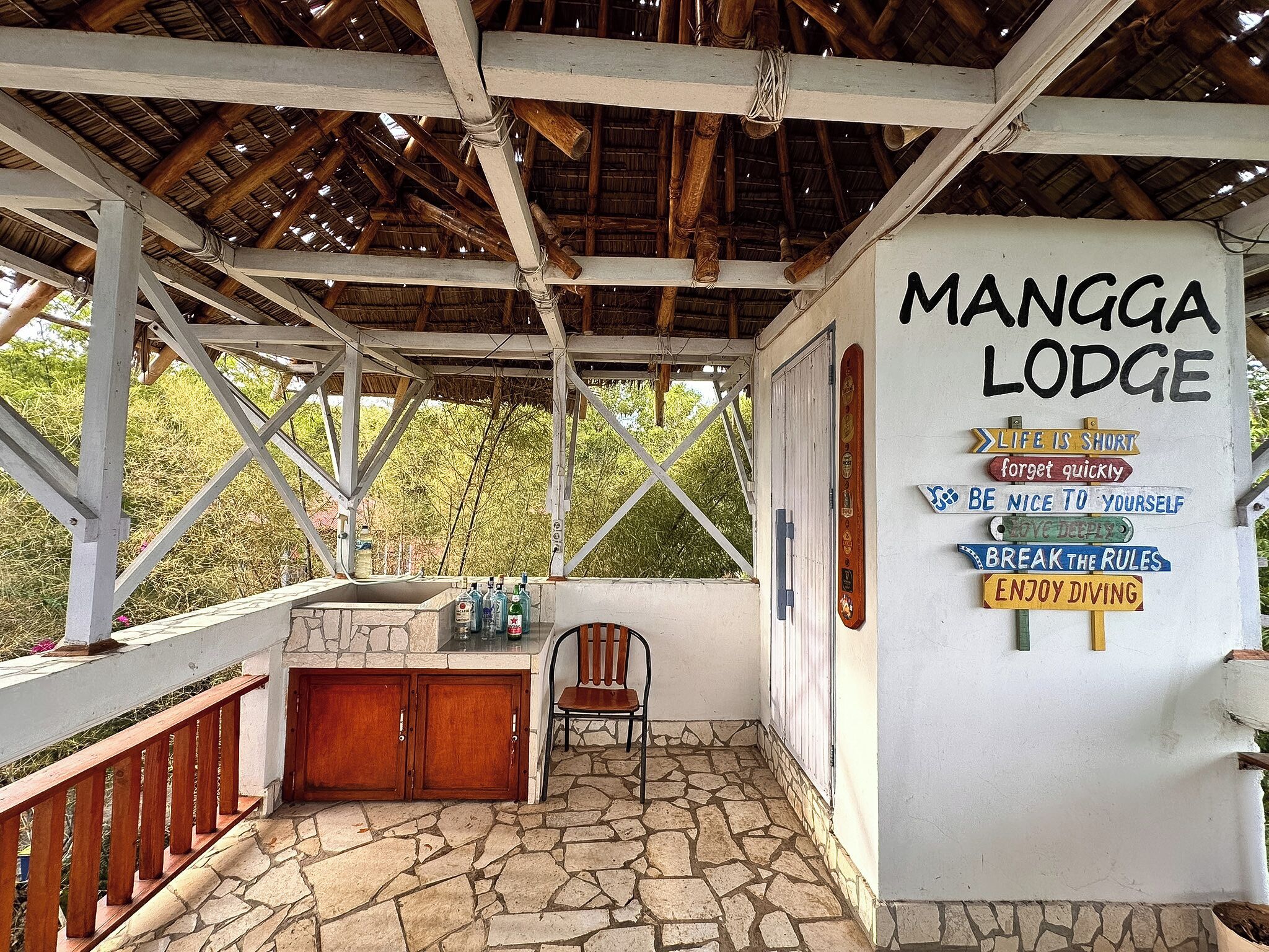 Un'area esterna coperta con pavimento in pietra, una panchina in legno e un cartello che riporta il nome 'Mangga Lodge' insieme a diverse frasi motivazionali.