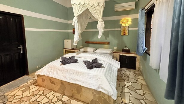 A bedroom with a bed, a nightstand, a wall-mounted air conditioner, and a window with curtains. - Mangga Lodge (Bira)