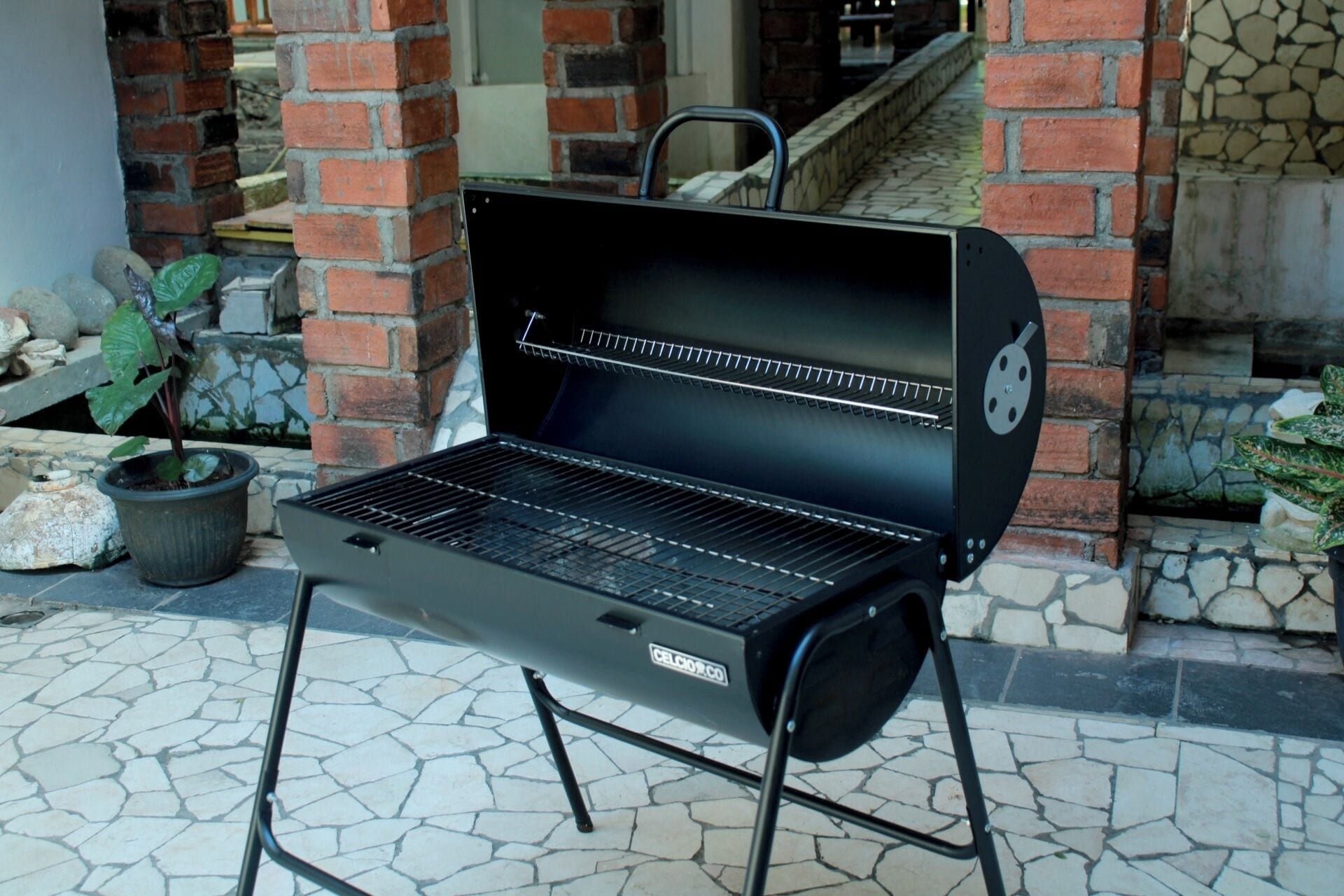 A black outdoor grill with a metal handle, positioned on a stone-tiled patio with a brick wall in the background.