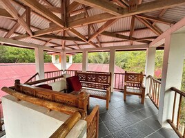 Uma área de estar ao ar livre coberta, com bancos e grades de madeira, cercada por uma construção com telhado vermelho.