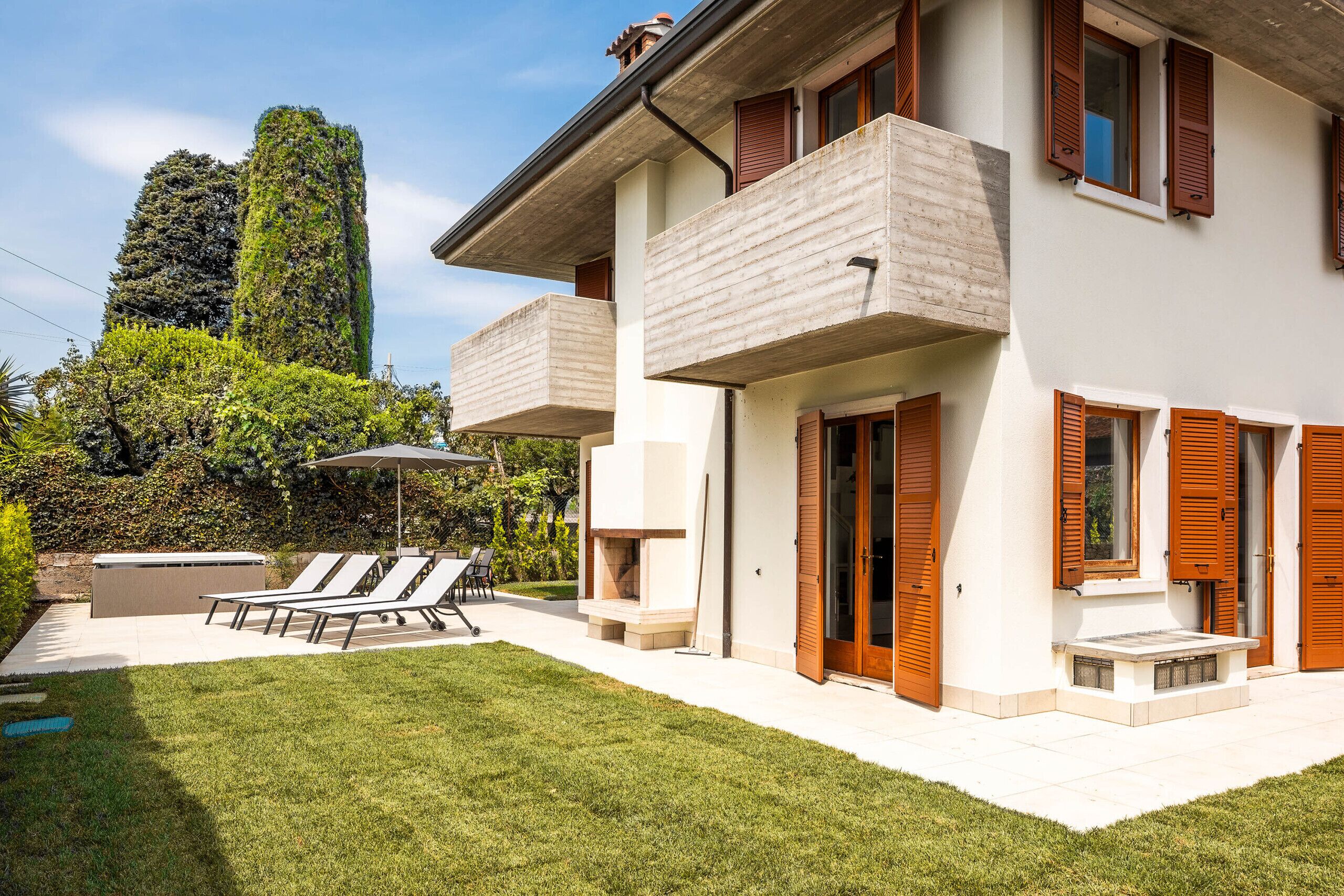 Una moderna casa a due piani con un balcone, un patio con poltrone relax e una piscina.