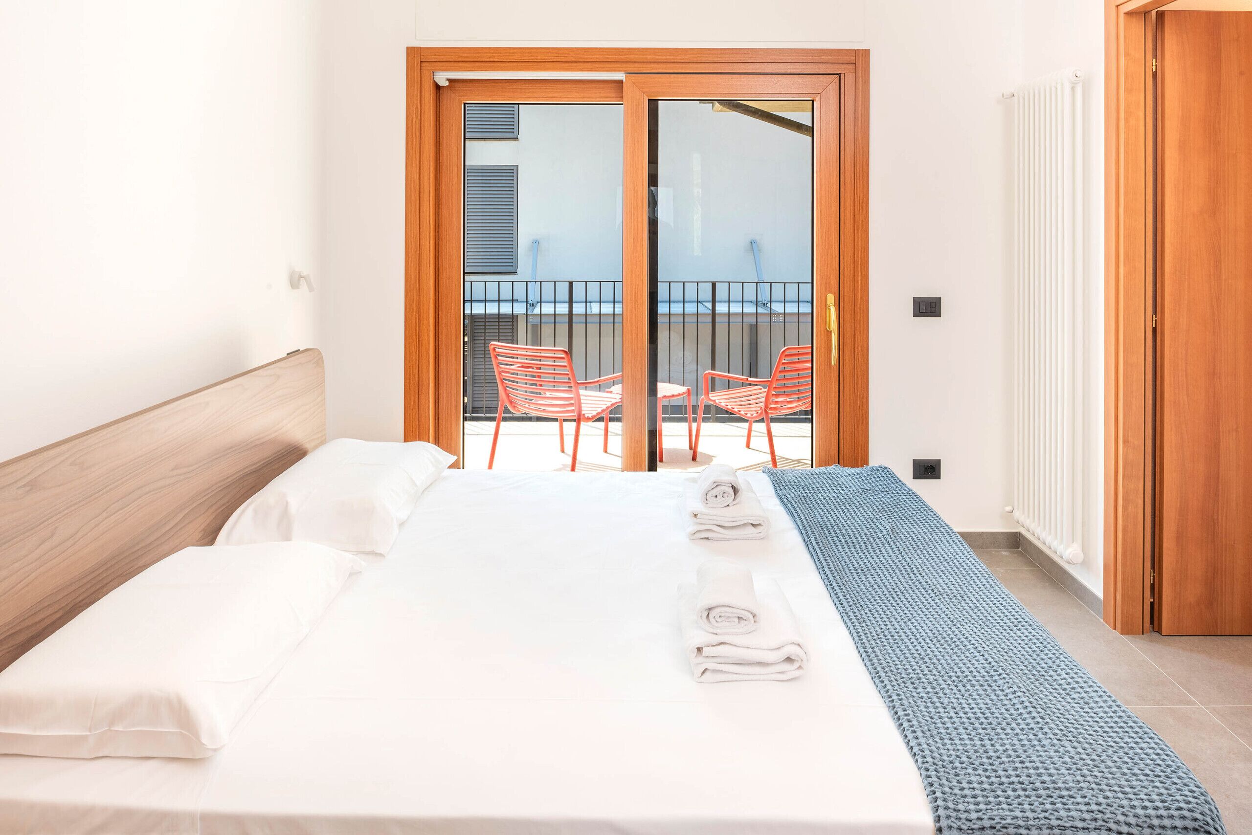 A hotel room with a bed, a window with blinds, a radiator, and a balcony with chairs.