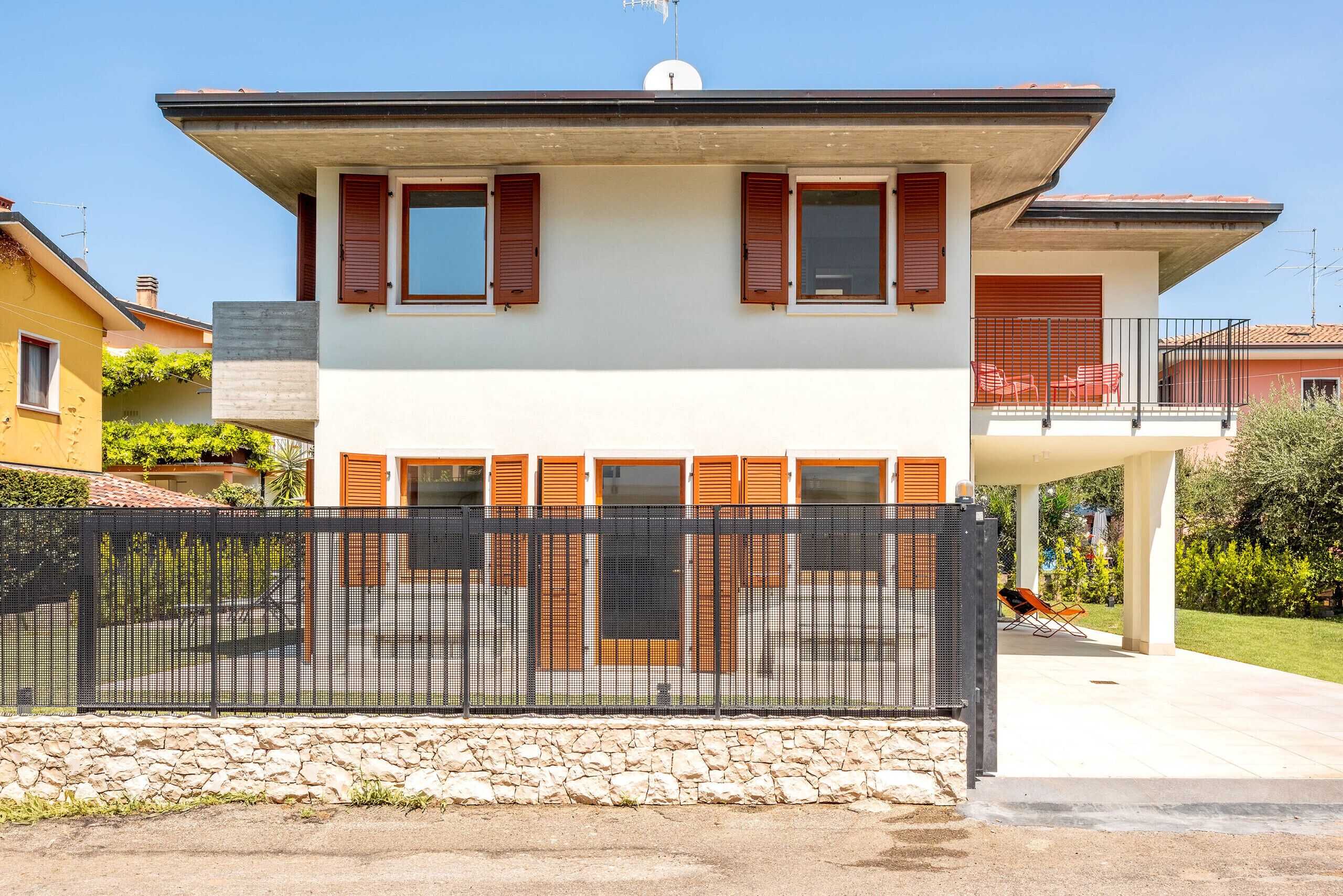 A modern two-story house with a balcony, a stone wall in front, and a metal fence.
