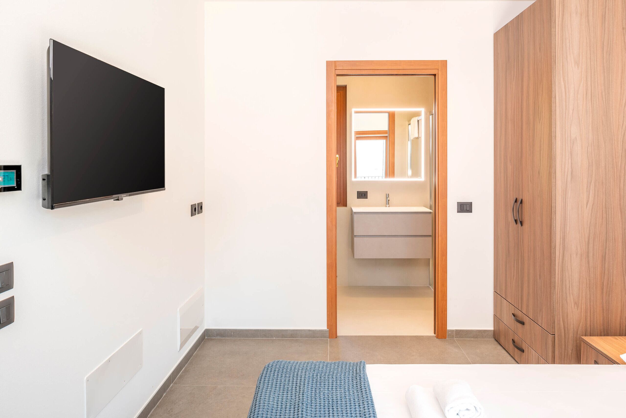 A hotel room with a flat-screen TV mounted on the wall, a wooden wardrobe, a bed with a blue textured blanket, and a bathroom with a mirror and sink.