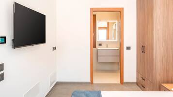 A hotel room with a flat-screen TV mounted on the wall, a wooden wardrobe, a bed with a blue textured blanket, and a bathroom with a mirror and sink.