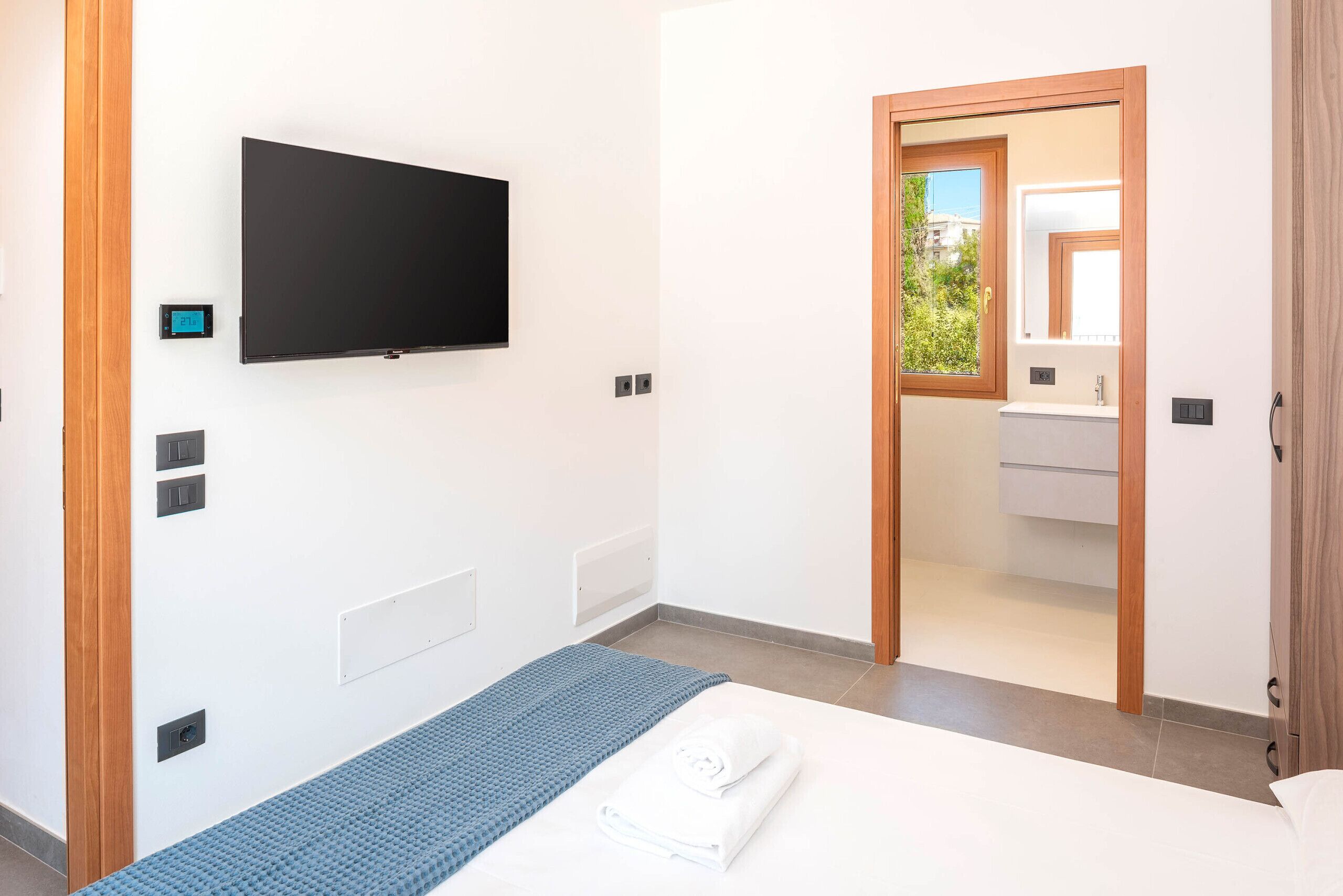Une chambre d’hôtel moderne dotée d’un téléviseur à écran plat fixé au mur, d’un lit avec un matelas bleu texturé, d’une serviette blanche et d’une vue sur la salle de bain à travers une porte en verre.