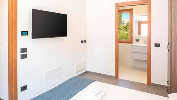 Une chambre d’hôtel moderne dotée d’un téléviseur à écran plat fixé au mur, d’un lit avec un matelas bleu texturé, d’une serviette blanche et d’une vue sur la salle de bain à travers une porte en verre.