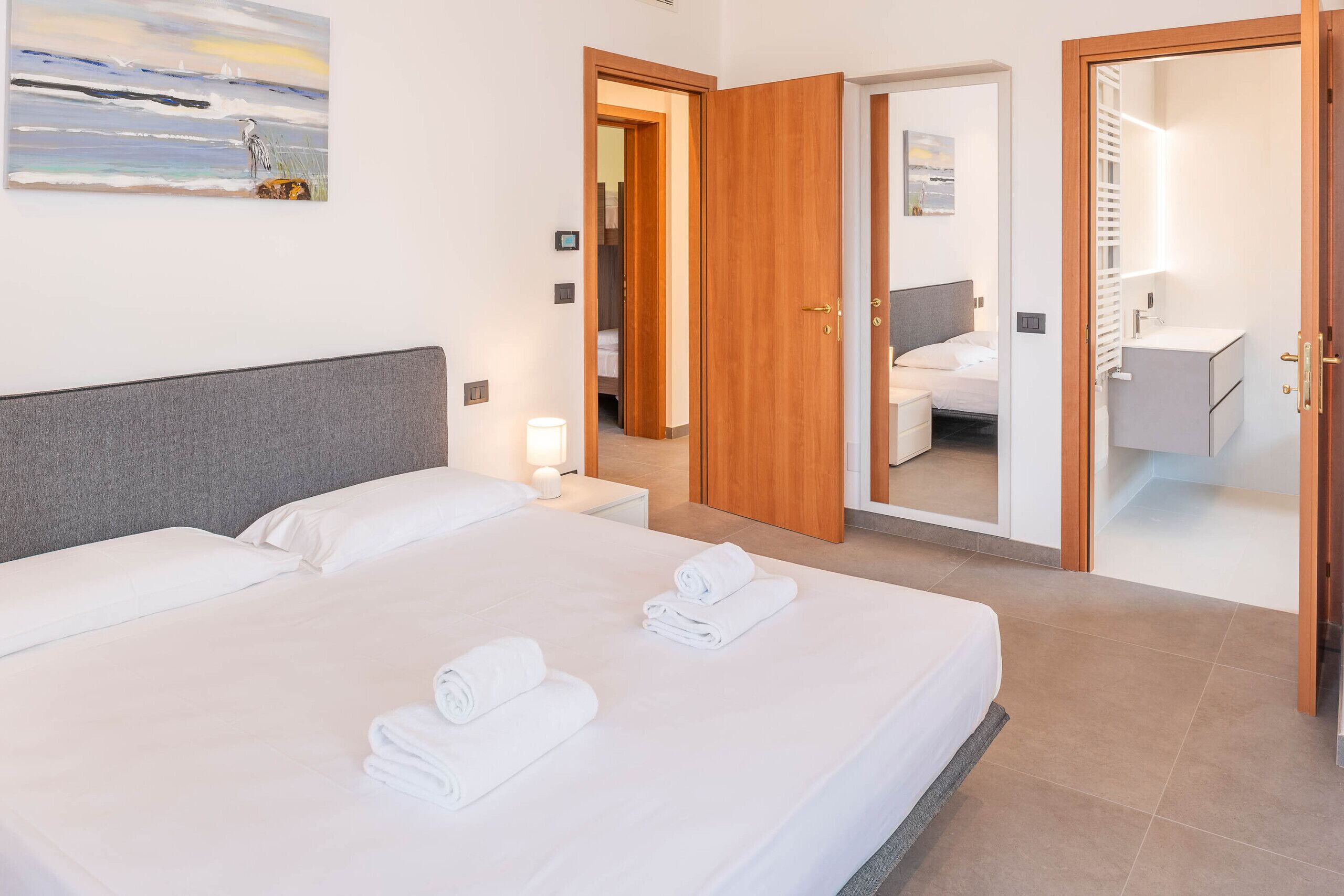 Une chambre d’hôtel avec un lit, une table de chevet, une lampe, un tableau au mur et une salle de bain visible à travers une porte ouverte.
