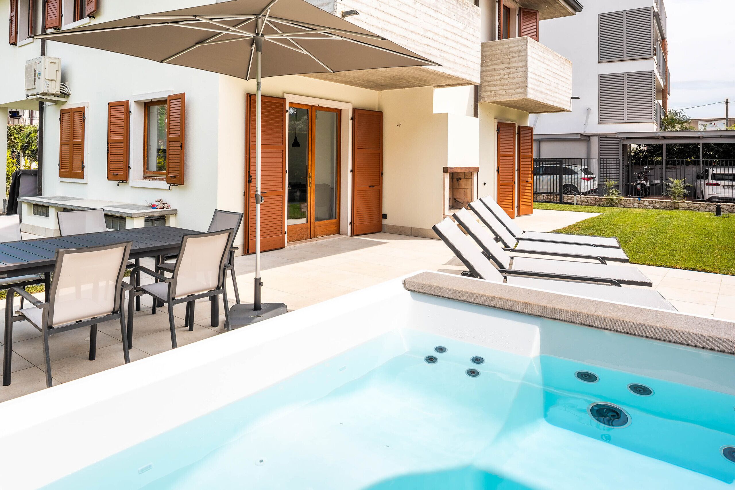 Un'area piscina moderna all'aperto con idromassaggio, un tavolo con sedie e un patio coperto con ombrellone.