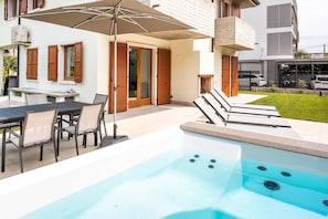 A modern outdoor pool area with a hot tub, a dining table with chairs, and a covered patio with an umbrella.