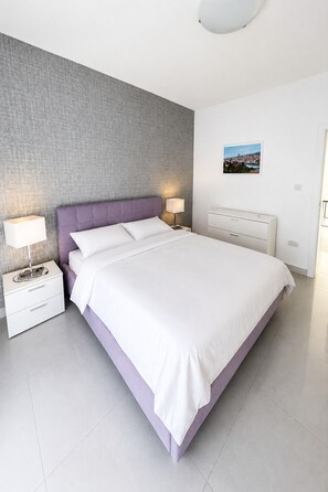 A modern bedroom with a large bed, two bedside tables with lamps, a grey textured wall, and a small picture on the wall. - Perfect 3BR Apartment with Coast View - Fl 1 (Naxxar)