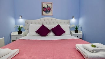 A neatly made bed with a red and white bedspread, two purple pillows, and a white headboard. There are two bedside tables with lamps, a framed floral artwork above the bed, and a small potted plant on each table.