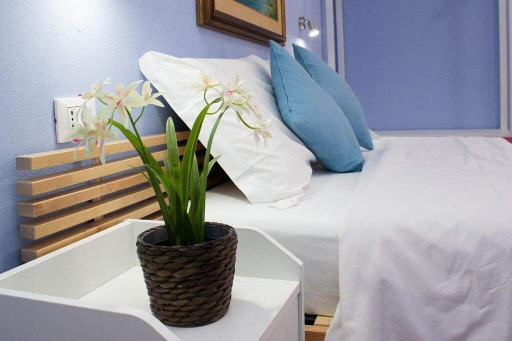 Een bed met witte beddengoed, een blauw kussen en een wit kussen. Er staat een gevlochten mand met een plantje op een witte nachtkastje.