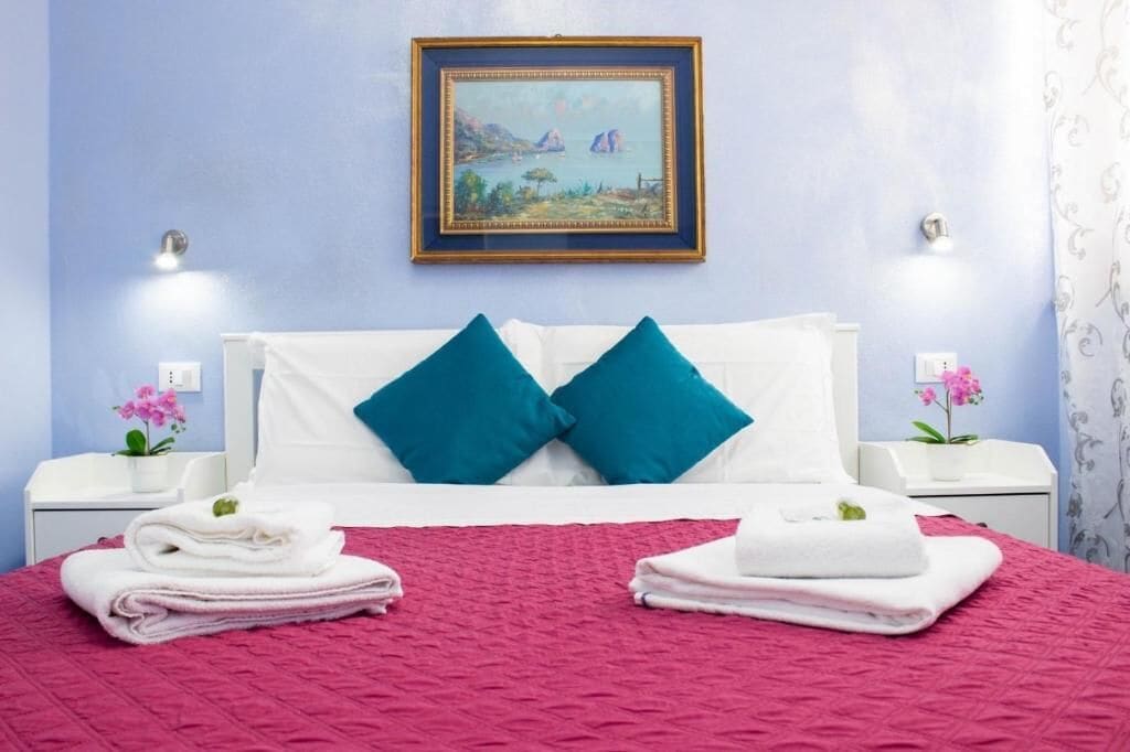 Una habitación de hotel con cama, almohadas, un cuadro en la pared y dos mesitas de noche con lámparas y flores.