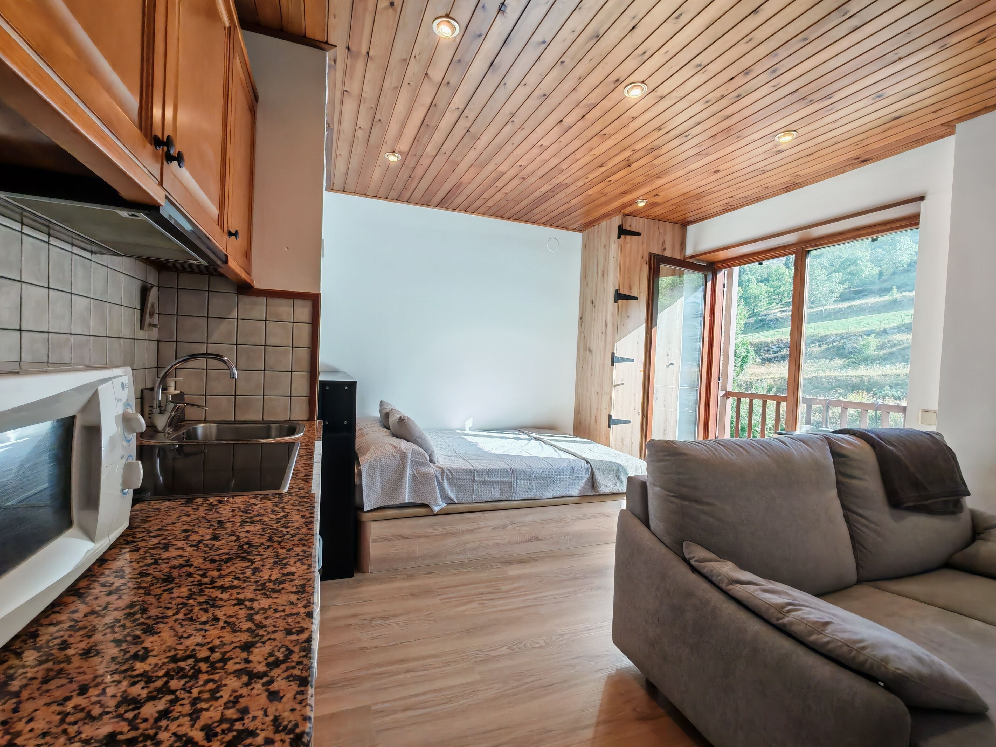 A modern kitchen with a microwave, sink, and wooden cabinets. There is a bed with a view of the outdoors through a large window.