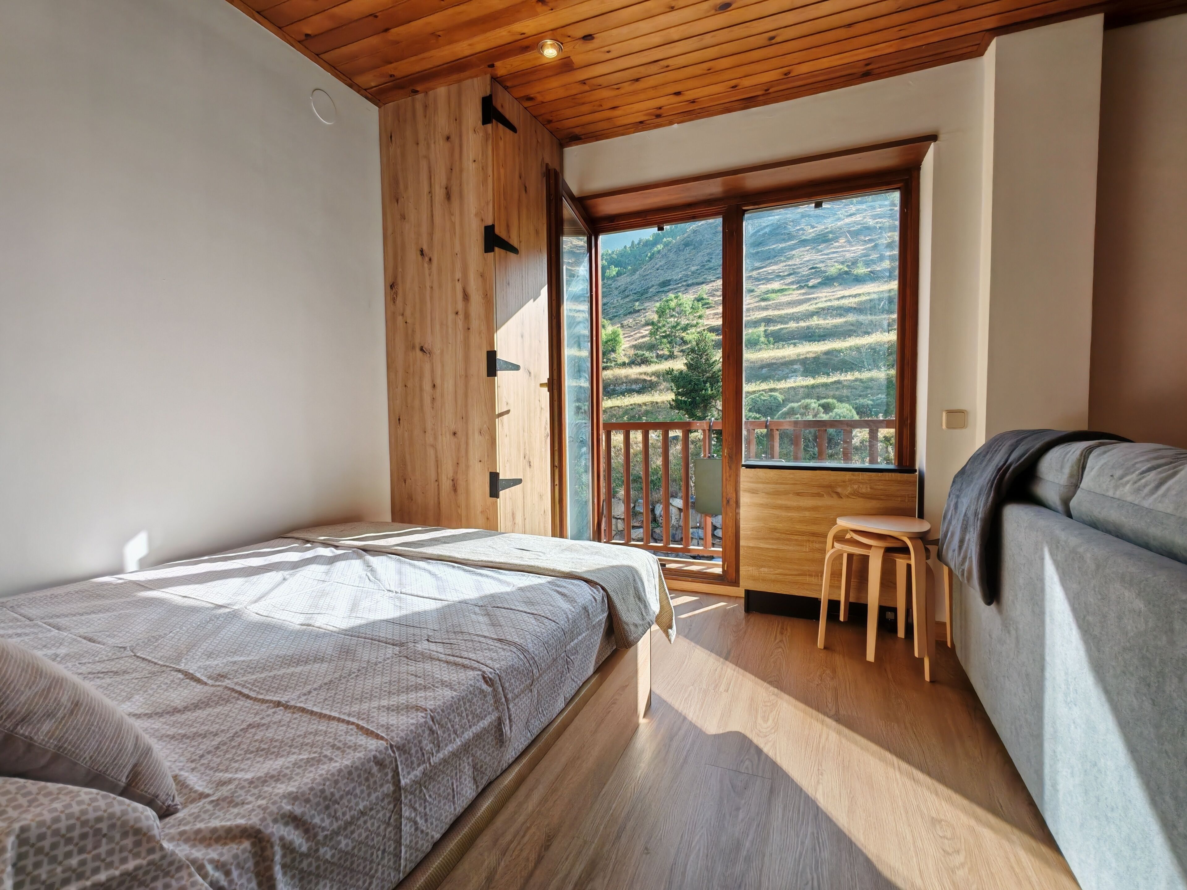 Ein Schlafzimmer mit Holzboden, einem Bett mit gemustertem Überwurf, einem kleinen Holzschemel und einem Balkon mit Blick auf eine hügelige Landschaft.