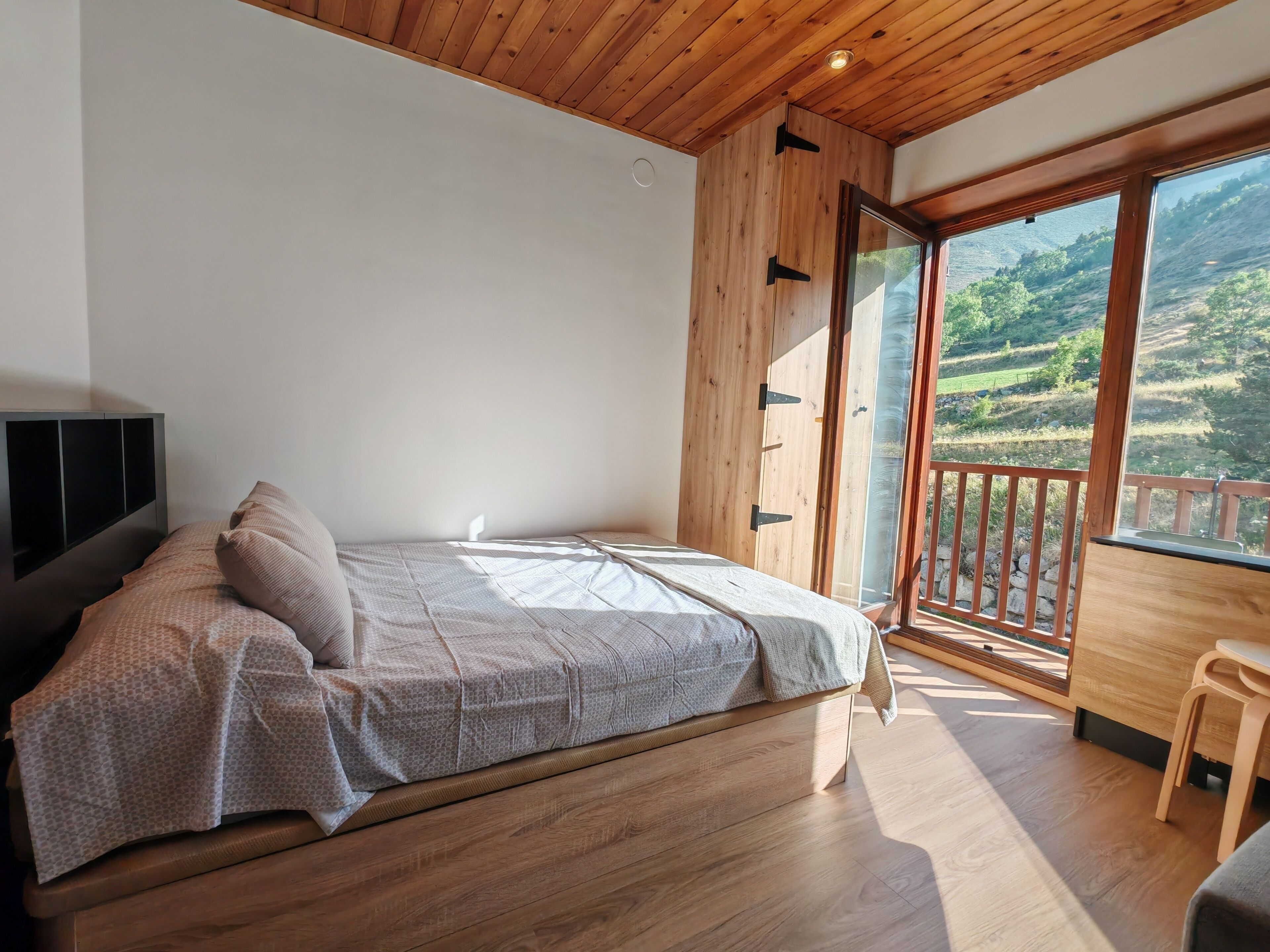 A bedroom with a bed, a TV, a wooden ceiling, and a balcony with a view of the countryside.