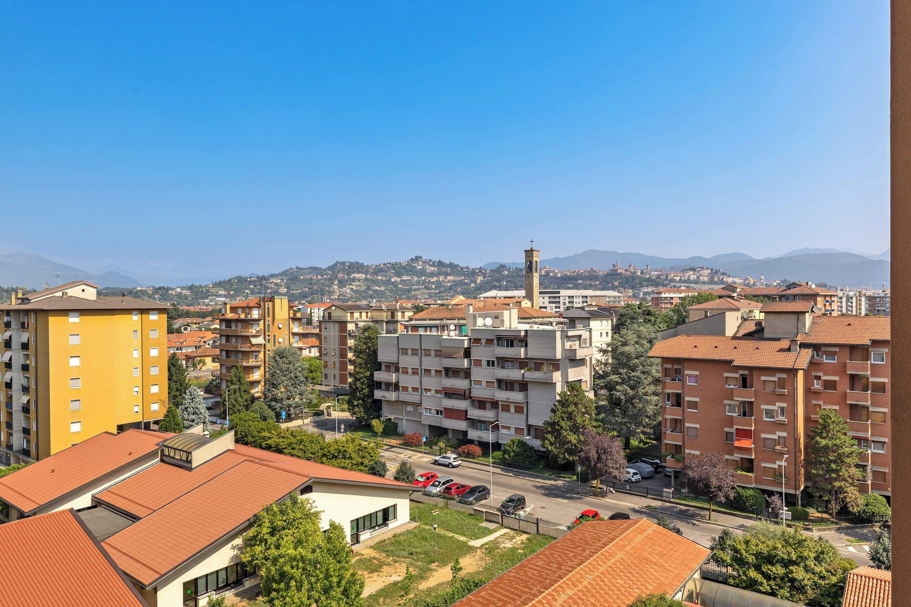 A residential area with multi-story buildings, a road with parked cars, and a distant view of a hill with a tower.