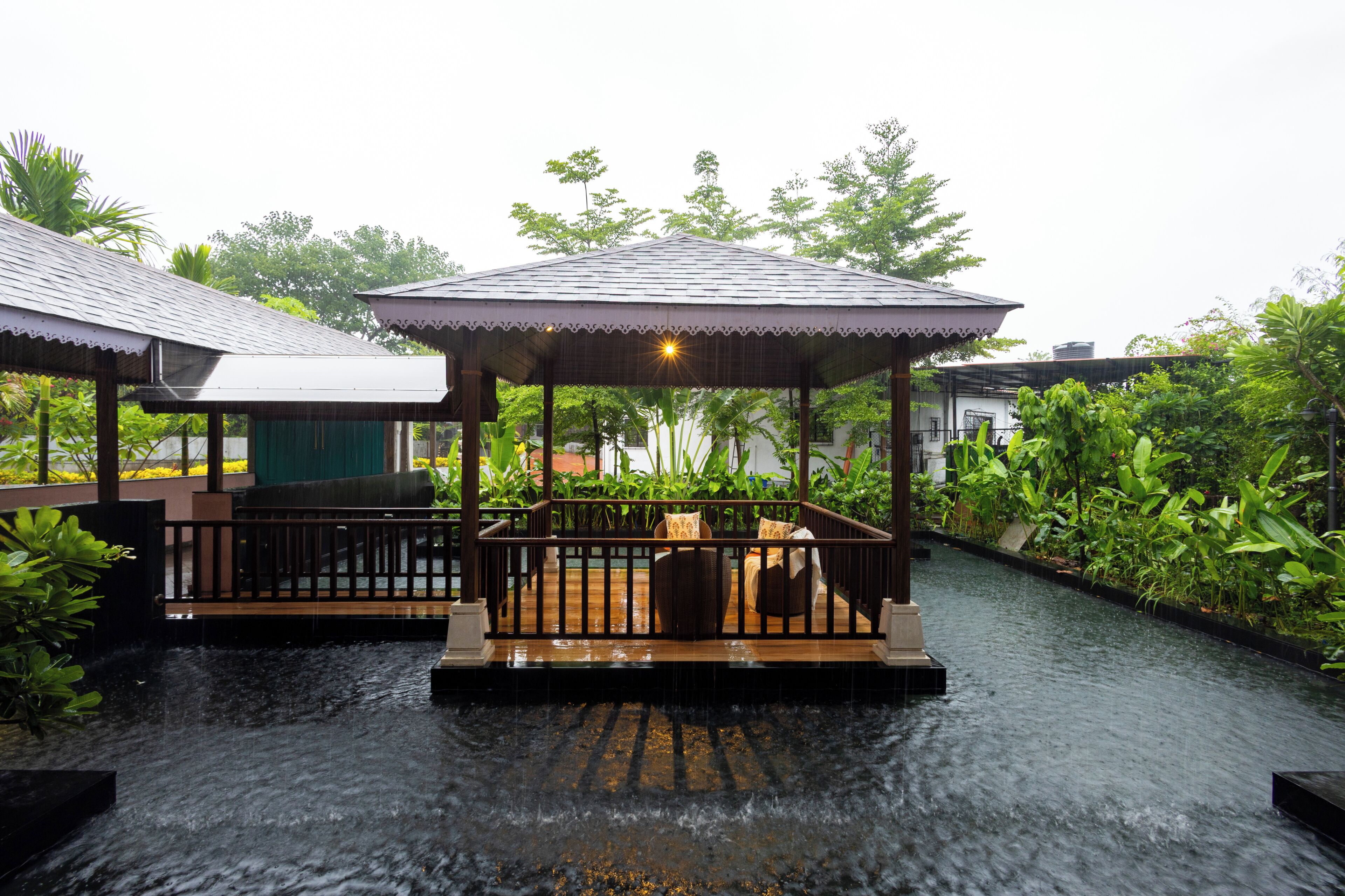 A covered outdoor seating area with a wooden deck and a canopy, surrounded by lush greenery and a water channel.