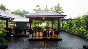 A covered outdoor seating area with a wooden deck and a canopy, surrounded by lush greenery and a water channel.