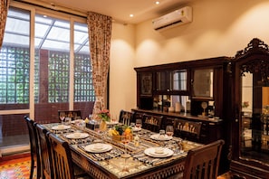 A dining room with a long table set for six, surrounded by chairs, with a large window and a decorative cabinet.