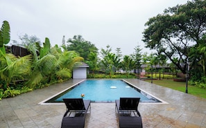 A swimming pool with two lounge chairs facing the water, surrounded by lush greenery and a small building in the background.