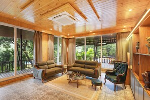 A spacious room with a wooden ceiling, large windows, a sofa set, a coffee table, and a decorative chair. - ELIVAAS Aaraa 4BHK Luxury Pool Villa (Karjat)