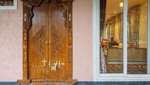 A wooden double door with intricate carvings, a window with patterned curtains, and a glimpse of a room with a sofa and a lamp.
