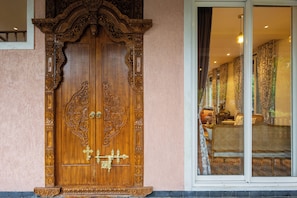 A wooden double door with intricate carvings, a window with patterned curtains, and a glimpse of a room with a sofa and a lamp.