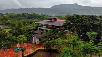 A two-story building with a balcony, surrounded by lush greenery and a view of a hill.