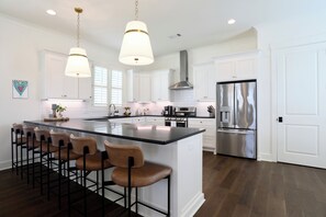 Une cuisine moderne dotée d’un grand îlot central, de tabourets de bar, d’appareils électroménagers en acier inoxydable et de luminaires suspendus.