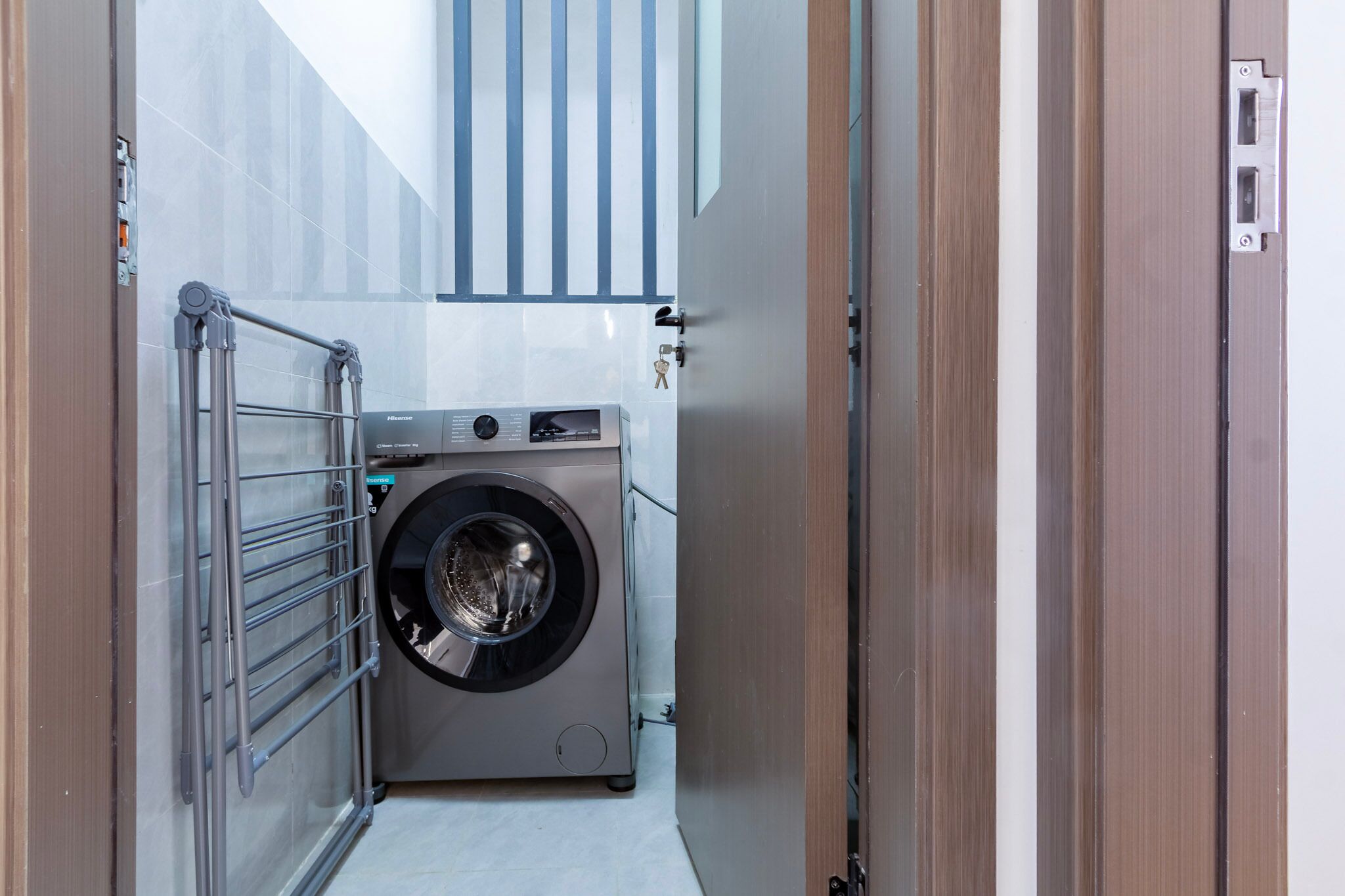 A compact laundry room with a front-loading washing machine, a metal drying rack, and a door with a key lock.