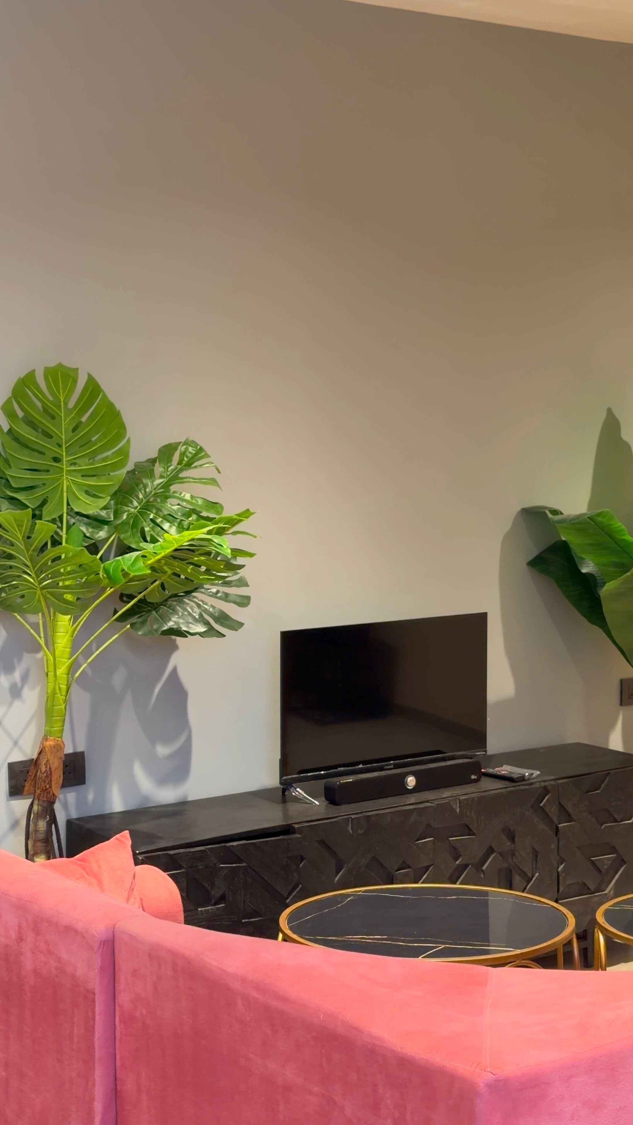 A room with a flat-screen TV mounted on a wall, a black entertainment unit with a gold circular table, and a large green plant.