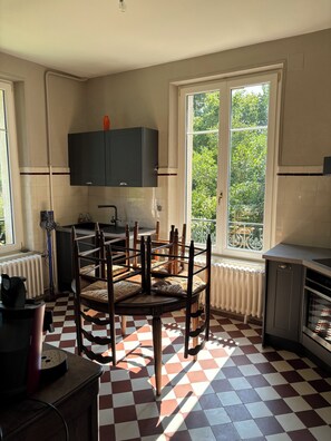 A kitchen with a checkered floor, a dining table with chairs, a window with a view of greenery, and a radiator. - Argonne Guesthouse (Dombasle-en-Argonne)