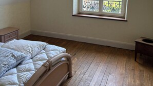 Un dormitorio con suelo de madera, una cama con ropa de cama blanca, una ventana que da a vistas de zonas verdes y edificios, y una mesita de noche pequeña.