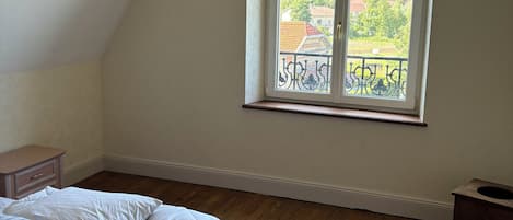 Un dormitorio con suelo de madera, una cama con ropa de cama blanca, una ventana que da a vistas de zonas verdes y edificios, y una mesita de noche pequeña.