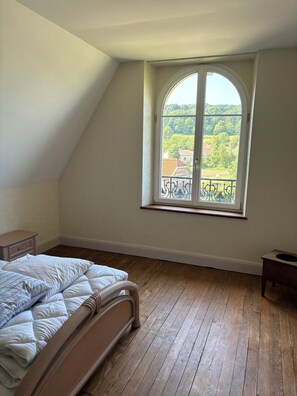 A bedroom with a wooden floor, a bed with white bedding, a window with a view of greenery and buildings, and a small bedside table. - Argonne Guesthouse (Dombasle-en-Argonne)