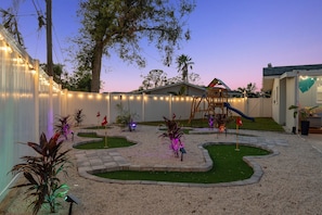 A backyard with a play area, including a slide and a swing set, surrounded by a white fence and string lights. - Family Home Retreat•Hot Tub•Heated Pool•Mini-Golf (Seminole)