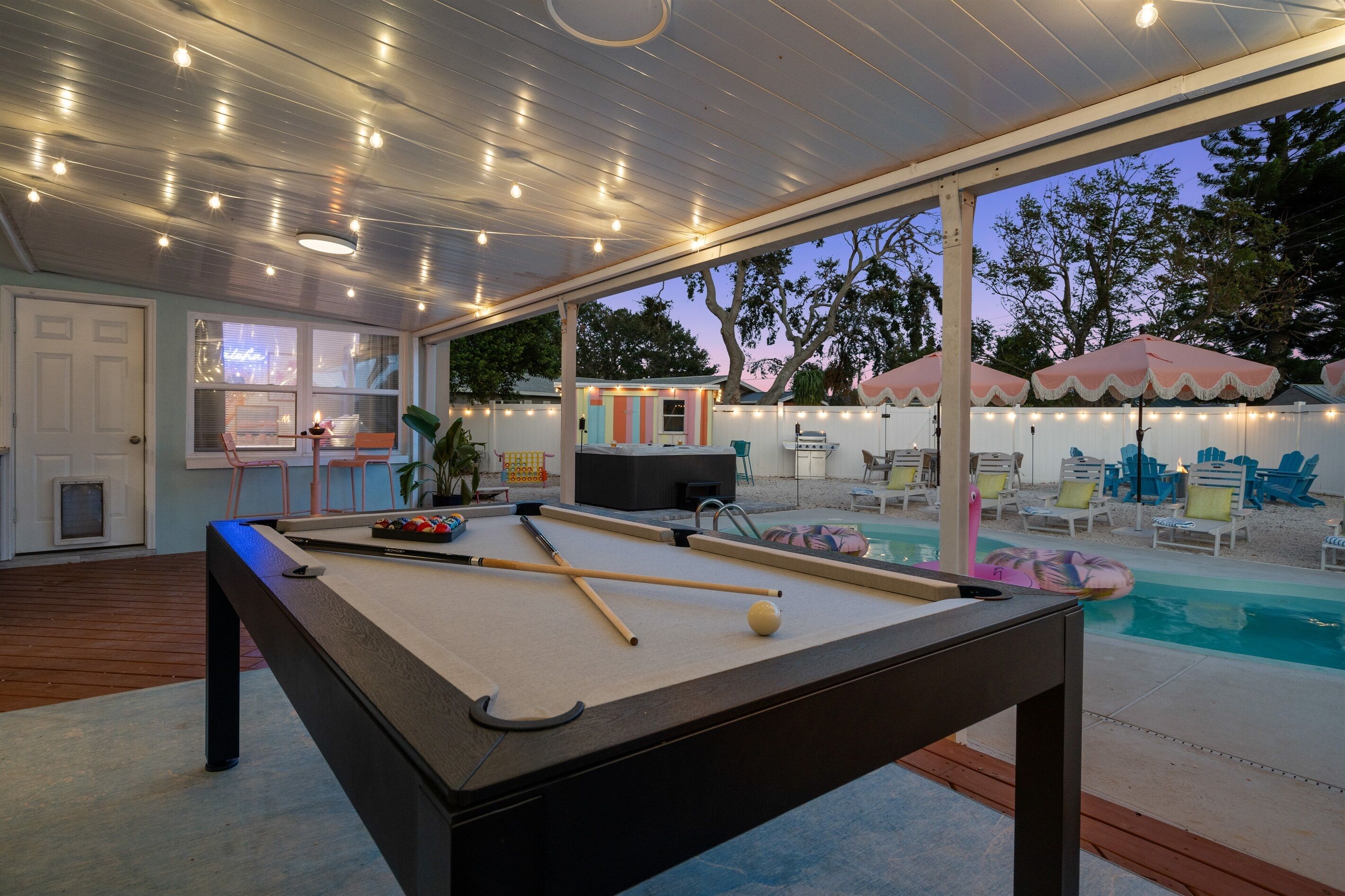 A pool table with pool cues and balls, a pool table in a covered area with string lights, a pool with lounge chairs and umbrellas, and a building with a colorful facade in the background.