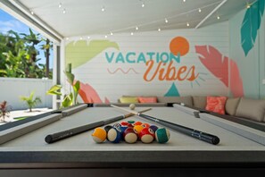 Ein Billardtisch mit Billardkugeln und Spielstöcken, ein Sofa mit Kissen und eine Wand mit einem 'VACATION Vibes'-Wandbild.