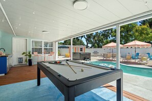 Ein Billardtisch in einer überdachten Außenbereich mit Blick auf ein Schwimmbad und Terrassenmöbel.