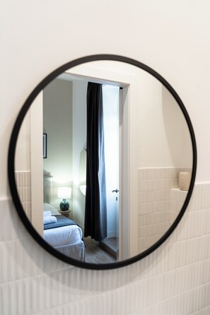 Un espejo circular refleja una habitación con una cama, una mesita de noche, una lámpara y un baño con un inodoro y un perchero para toallas.