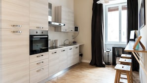 Una cucina moderna con elettrodomestici in acciaio inossidabile, forno integrato e microonde. Si vede una finestra con tende, un tavolino con un bollitore e uno sgabello.