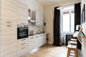 A modern kitchen with stainless steel appliances, a built-in oven, and a microwave. There is a window with curtains, a small table with a kettle, and a stool. - Charming Guesthouse in a Central Location Comfortable Stay for Couple & Families (Roma)