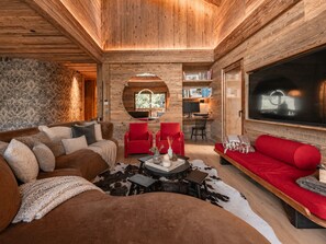 Ein gemütliches Blockhauss-Interieur mit Holzbalkendecke, einem großen Fernseher, einem Kamin und einer Sitzecke mit einem roten Sofa und einem braunen Schlafsofa.